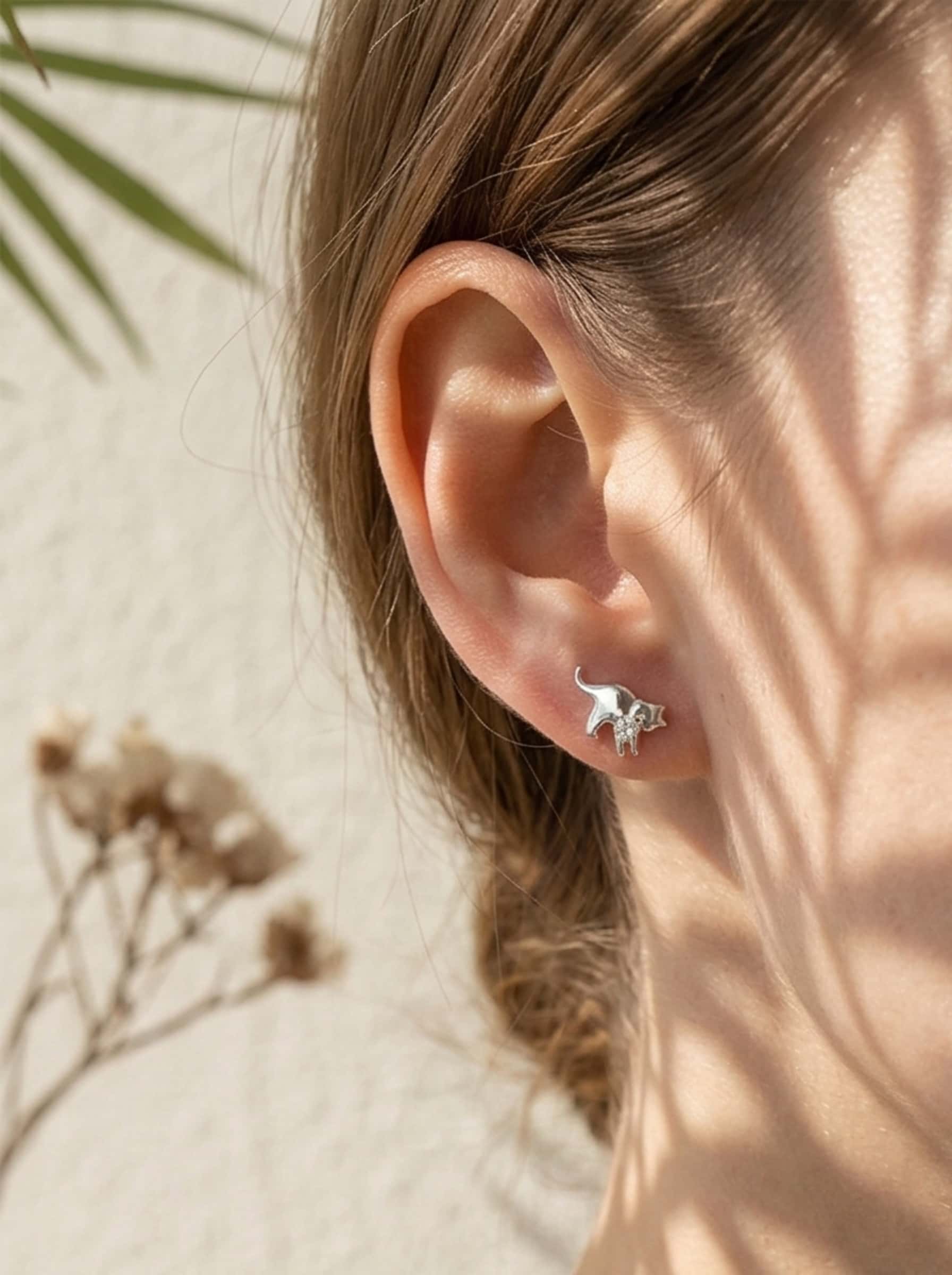 A woman modeling elegant sterling silver cat earrings outdoors in warm sunlight with artistic palm leaf shadows
