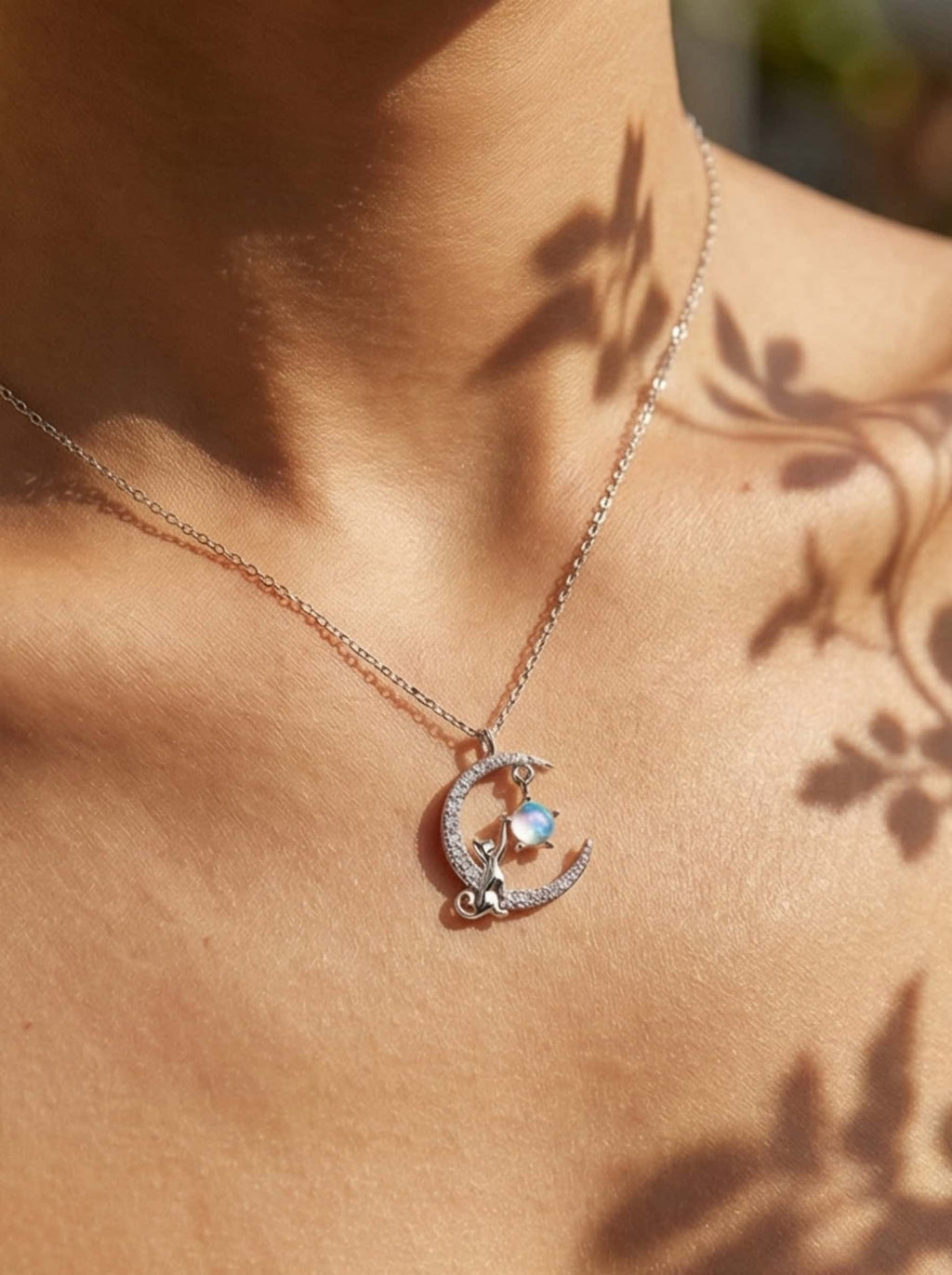 Close-up of a cat pendant necklace worn on the collarbone, showcasing a silver feline reaching for a luminous moonstone under dappled sunlight.