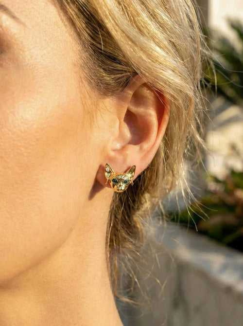 Close-up of a person wearing a gold sphinx-cat earrings outdoors.